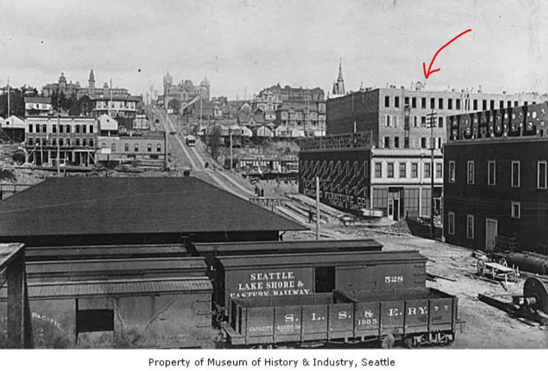 The Grand Hotel's back has an arrow pointed to it. Construction is underway on the street and many buildings. A cable car heads up Madison Street towards the Central School. The Rainier Hotel can be seen in the distance. Railroad boxcars and flat cars are in the foreground.
