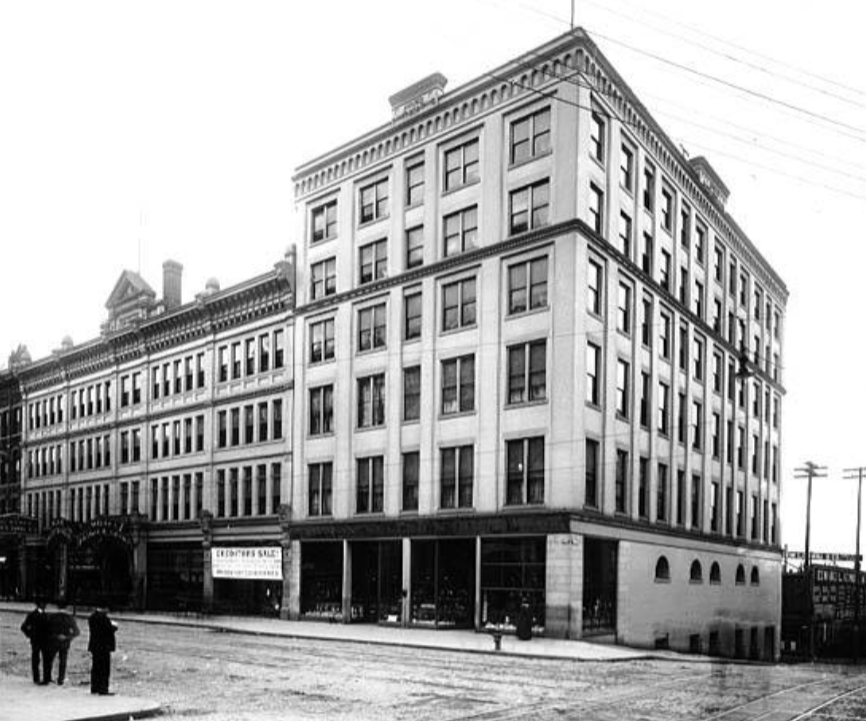 Photograph of the Rainier-Grand Hotel, seen looking southwest from the intersection of First and Madison. The new annex is visible on the opposite corner from the photographer. A sign over a first floor window says "Christmas Sale".
