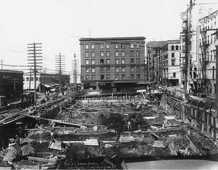 Photograph of the onset of construction on a block-long, half-block wide site between Madison, Marion, Western, and Post. The foundation is being laid for a warehouse. The site was considered for expansion of the Rainier-Grand Hotel. 