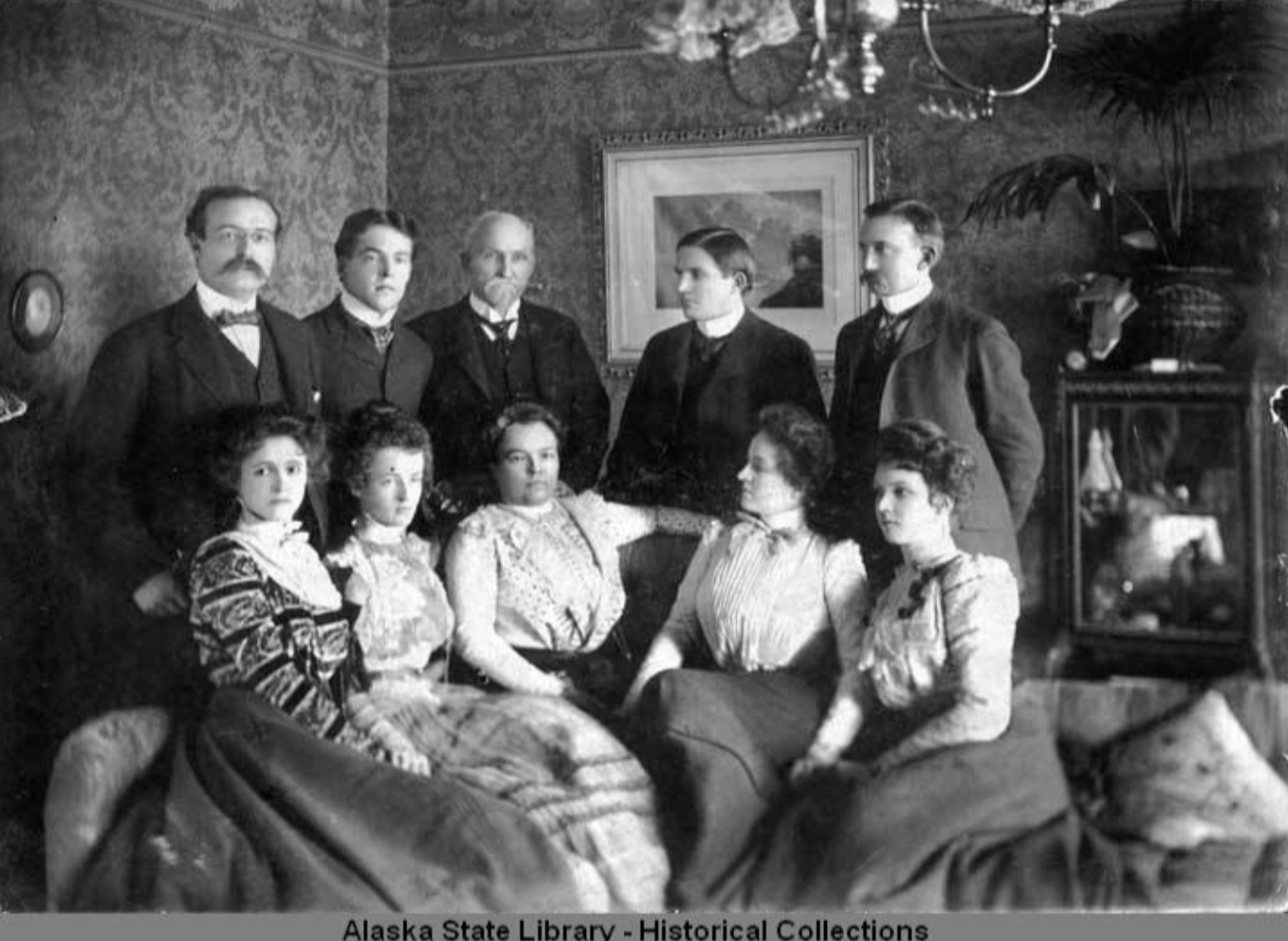 Photograph of members of the Noyes Family. Five men in suits stand behind five women in dresses sitting on couches and chairs. John Noyes with his long white beard is recognizable standing in the middle, presumably with his wife Elmira in front of him. The other four couples are their children and spouses. All are white.