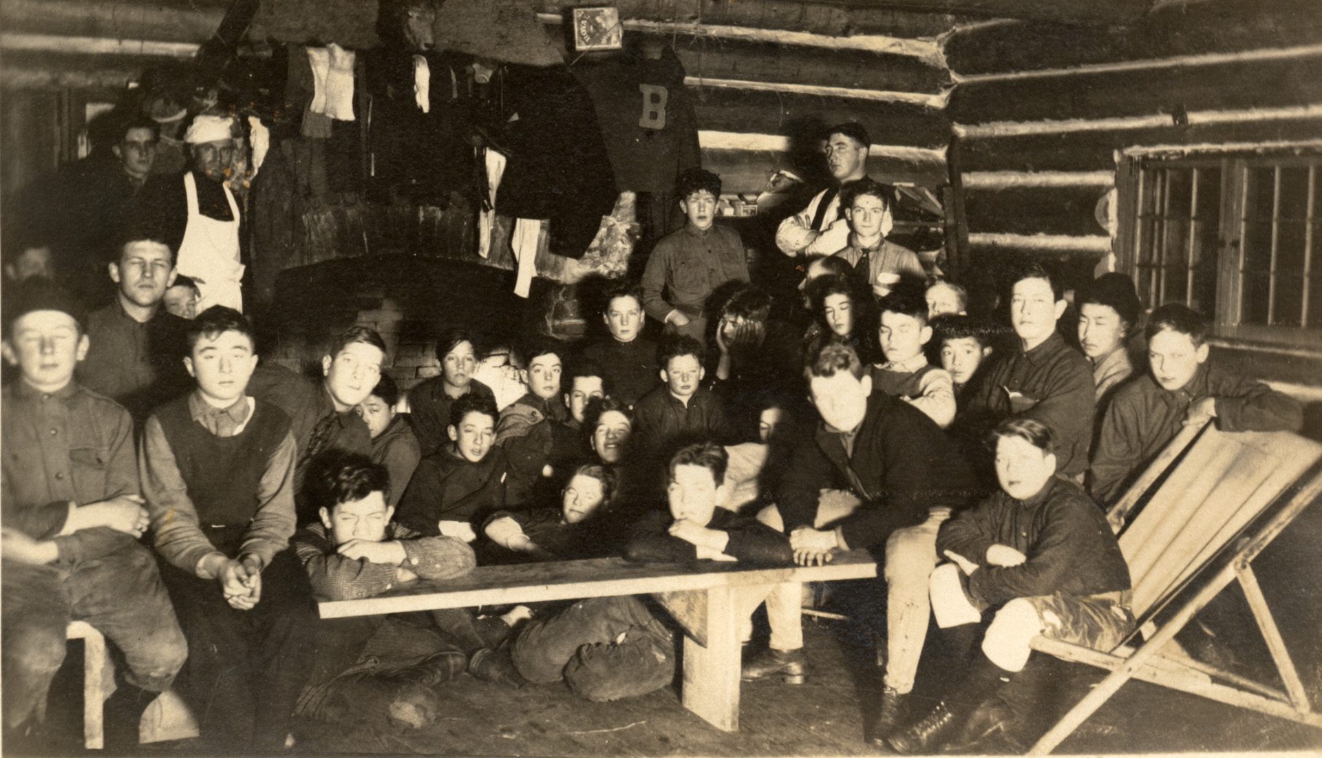 Youth in sweaters crowd in front of a large fireplace, sitting on anything. Clothes hang over the fire drying. Several leaders stand in the back, including one in apron and chef's hat. George Nakashima is on the far right.