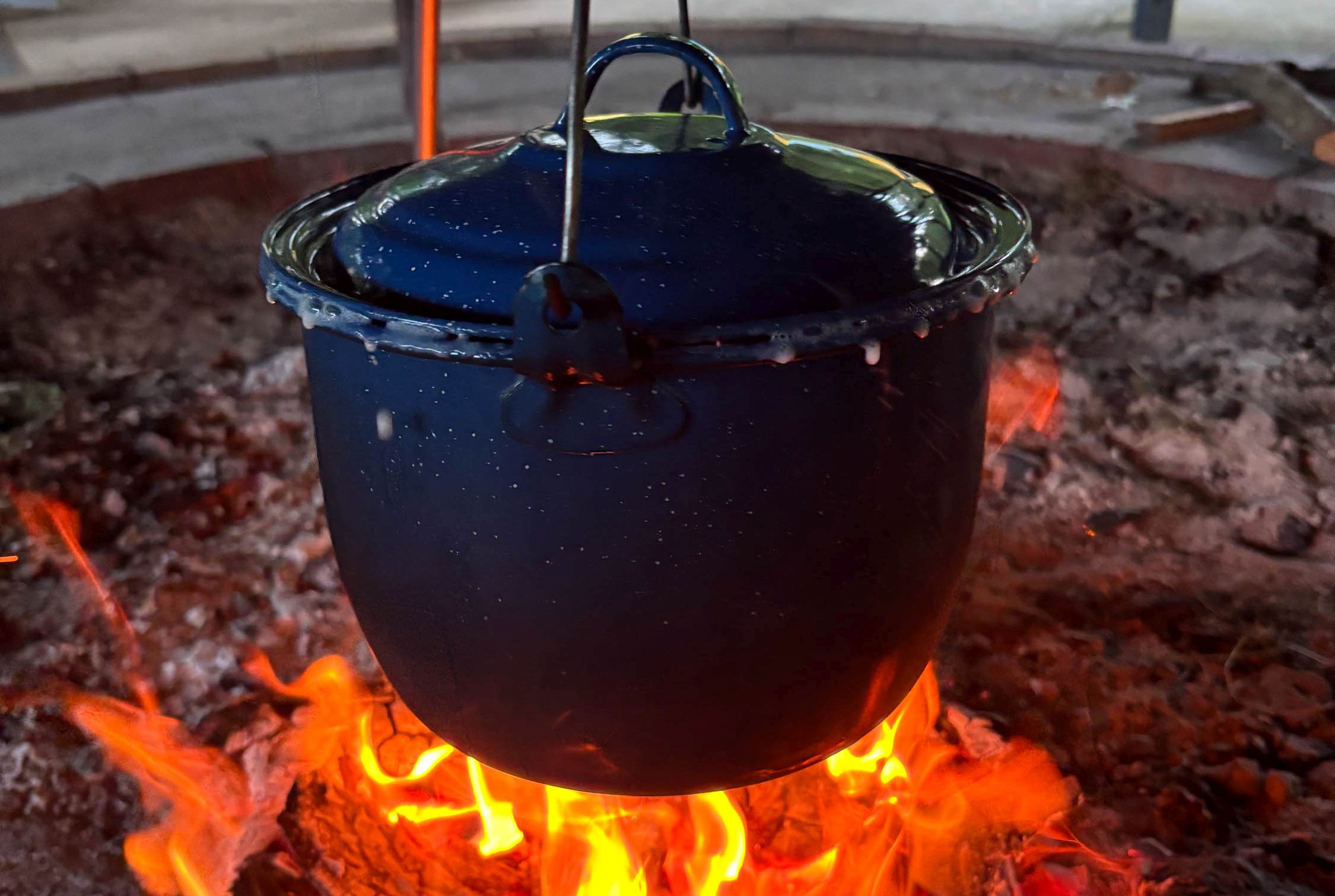 A pot hangs over a red fire, with it lid lifted as the contents boil over.